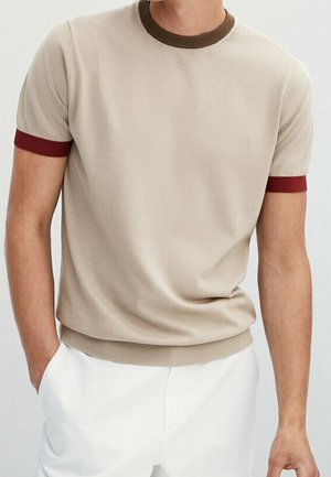 Mann trägt einen beigefarbenen Kurzarm-Pullover mit braunem Kragen und roten Ärmelbündchen, kombiniert mit weißen Hosen, Hände in den Taschen.