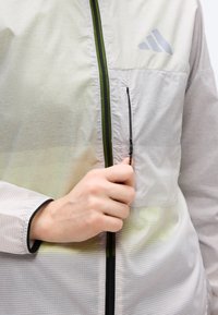 Το χέρι ενός ατόμου τραβά το φερμουάρ ενός ανοιχτόγριζου μπουφάν Adidas με κάθετη τσέπη στο στήθος και ορατό λογότυπο της μάρκας.