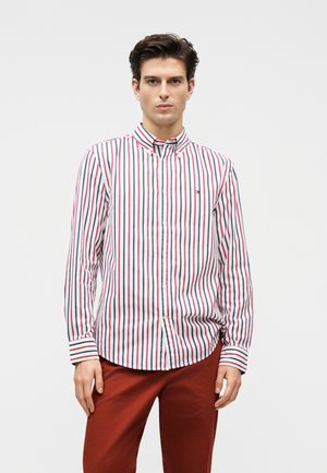 Homme face à l'avant portant une chemise blanche boutonnée avec des rayures verticales rouges, bleues et noires et un pantalon rouge sur un fond clair uni.