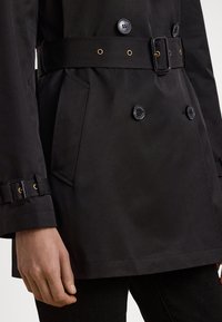 Czarny płaszcz trencz z dwurzędowym zapięciem na guziki, paskiem i bocznymi kieszeniami. Charakteryzuje się gładką fakturą oraz złotymi akcentami na guzikach i pasku.