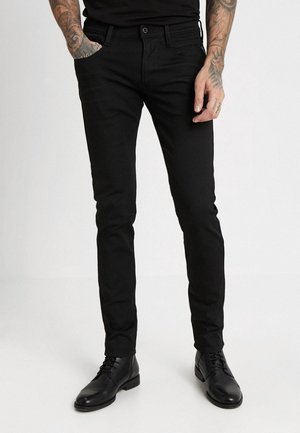 Homme portant un jean noir slim et des bottes habillées en cuir noir, avec des bras tatoués et une main dans la poche, debout sur un fond blanc uni.