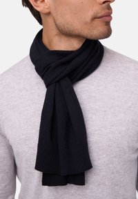 Écharpe en tricot noir drapée autour du cou, avec une texture côtelée et des bords nets, portée sur un pull à col rond gris clair.