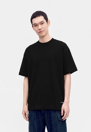 Giovane uomo che indossa una t-shirt nera semplice oversize e jeans cargo blu scuro in piedi davanti a uno sfondo chiaro uniforme.