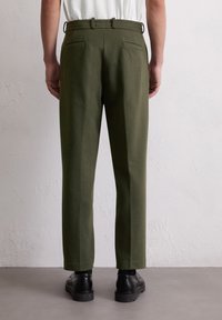 Pantalon vert olive à coupe droite, doté de deux poches arrière, de passants de ceinture et d'une texture lisse, associé à des chaussures noires.