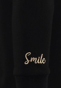 Tejido de sudadera negra con puños acanalados que presenta el texto bordado en dorado "Sonríe" en la manga izquierda. Textura suave con un corte ajustado.