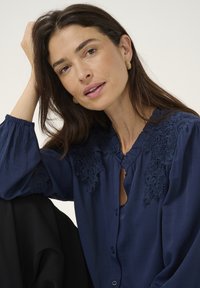 Blouse marine avec des détails en dentelle complexes sur les épaules, tissu doux, fermeture à boutons et manches bouffantes froncées. Tissu noir en arrière-plan.