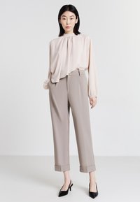 Blusa con drappeggio frontale, colore beige chiaro; abbinata a pantaloni grigi chiari su misura con orli risvoltati; modella che indossa tacchi a spillo neri.