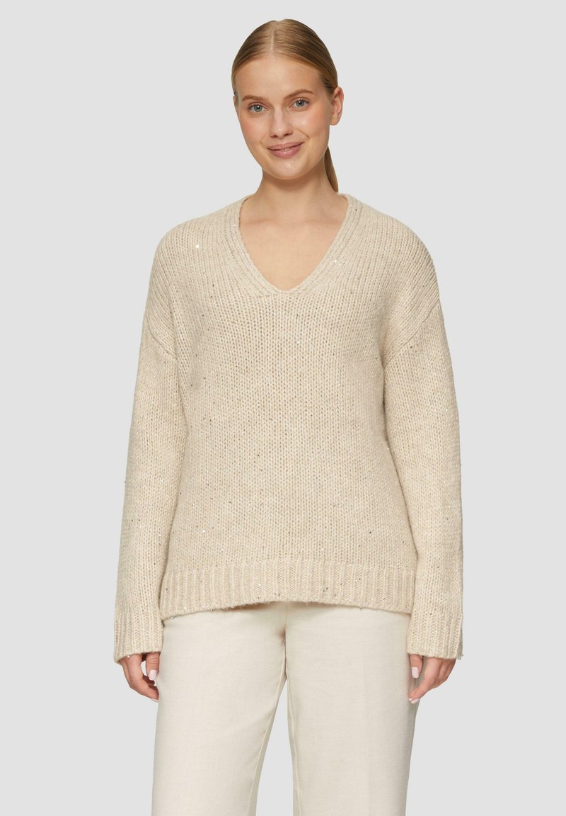 Beige oversized trui met een V-hals, ribgebreide zoom en mouwen, gestructureerd breiwerk en subtiele sparkles. Ggedragen met lichtgekleurde broeken.