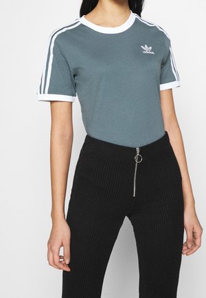 Kobieta w zielonej koszulce Adidas z białymi paskami na rękawach oraz czarnych prążkowanych spodniach z przednim zamkiem błyskawicznym i pierścieniowym uchwytem.