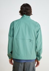 Light green, lightweight jacket with a high collar, elastic cuffs, and a back pocket. Smooth texture, simple design, and relaxed fit.