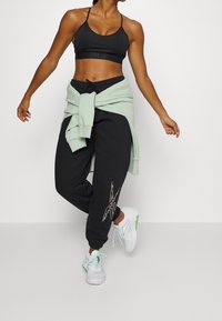 Černá sportovní podprsenka, volné černé tepláky s grafickým logem a mátově zelená mikina uvázaná v pase. Bílé tenisky se zelenými akcenty.