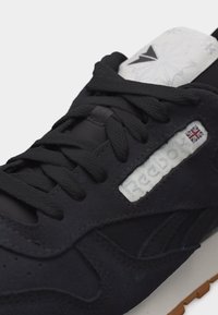 Μαύρο αθλητικό παπούτσι Reebok με μαύρα κορδόνια, λευκή ετικέτα με το λογότυπο που απεικονίζει τη σημαία του Ηνωμένου Βασιλείου και γκρι γλώσσα με έμβλημα αστέρι.