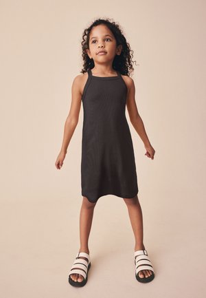 Giovane ragazza con capelli ricci che indossa un vestito nero senza maniche e sandali bianchi, in piedi con sicurezza davanti a uno sfondo beige uniforme.