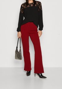Haut noir à manches longues avec des détails en dentelle, associé à un pantalon rouge taille haute et des escarpins noirs à bout pointu. Accessoirisé avec un sac métallisé.