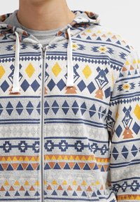 Felpa con cappuccio e zip di colore grigio chiaro, con motivi geometrici in blu navy, giallo e arancione. Tessuto testurizzato con cordini.