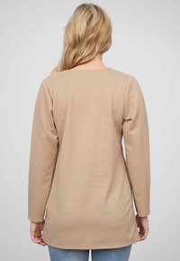 Beige dlouhý rukáv top s ribbanou texturou, kulatým výstřihem a zakřiveným lemem. Nosit přes modré džíny, viděno ze zadu.