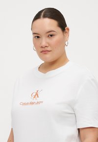 Bijela pamučna majica s kratkim rukavima koja ima narančasti logotip Calvin Klein Jeans, okrugli izrez. Glatka tekstura i opušteni kroj.