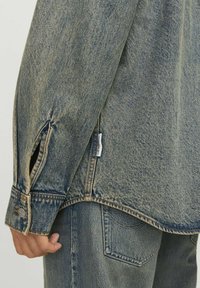 Camicia in denim con vestibilità rilassata, lavaggio azzurro chiaro, polsini con bottone e spacchi laterali. Tessuto testurizzato con dettagli di cucitura visibili.