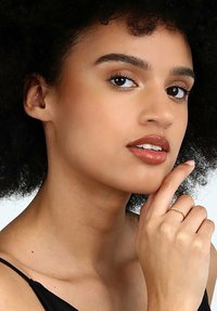 En ung person med lockigt svart hår, klädd i en svart topp, poserar med en hand på hakan och visar upp en subtil guldring på ett finger.