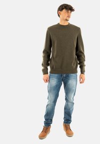 Olijfgroen gebreid sweater met een ronde hals, gedragen met lichtblauwe vervaagde jeans en bruine veterschoenen. Soepele textuur en slim fit.