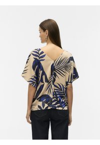 Beige top met een V-hals aan de achterkant, versierd met grote blauwe tropische bladeren, korte mouwen en een losse pasvorm. Gepaard met donkere denim jeans.