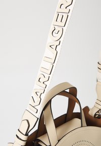 Beige leren handtas met zwarte accenten, geperforeerd cirkelontwerp, en een witte schouderband met "KARL LAGERFELD" in vette letters.