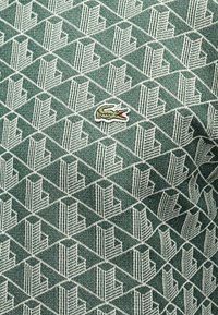 Πράσινο ύφασμα με επαναλαμβανόμενο λευκό γεωμετρικό μοτίβο και μικρό κεντητό πράσινο λογότυπο κροκόδειλου με κόκκινο στόμα στο κέντρο.