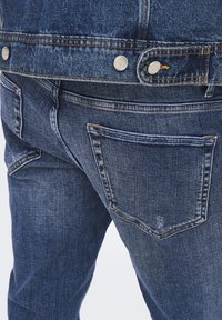 Jeans in denim blu scuro con una leggera texture sbiadita. Tasche posteriori standard con cuciture visibili e dettagli in metallo sui bottoni in vita.