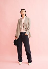 Striped blazer in beige and brown over a light pink shirt, paired with dark jeans and white pointed-toe heels. Black cap in hand.