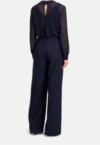 Marinblå jumpsuit med genomskinliga långa ärmar, nyckelhålsdetalj på ryggen och vida byxor. Det släta tyget och de knäppta manschetterna förbättrar designen.