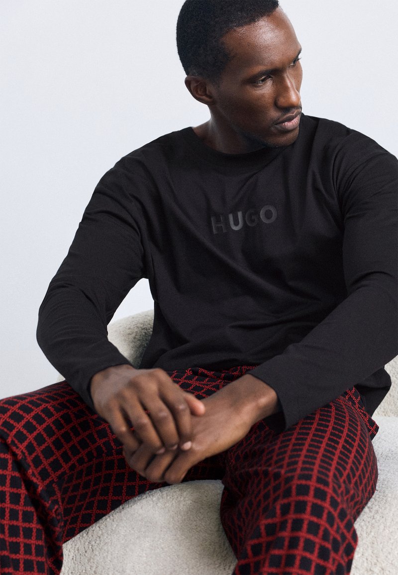 Schwarzes Langarmshirt mit "HUGO"-Logo und roten gemusterten Hosen mit Karodesign. Modell sitzt auf einer hellen strukturierten Oberfläche.