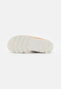 Sneaker med en tekstureret gylden overdel og hvid gummisål. Sålen har et bølgemønster og brandlogo indpræget nær hælen.