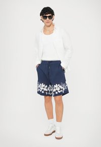 Shorts bleu marine à imprimé floral blanc, taille à cordon, associés à un débardeur blanc et une chemise blanche légère. Portés avec des lunettes de soleil et des baskets.
