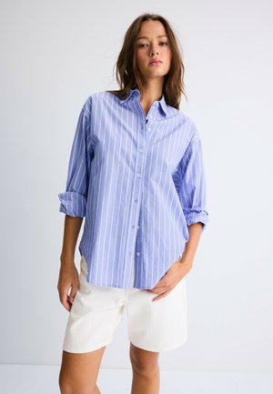Jeune femme portant une chemise à rayures bleu et blanc avec boutons, manches retroussées, et un short blanc, debout devant un fond clair uni.