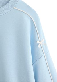 Felpa azzurra chiara con colletto a costine, piping bianco lungo le cuciture delle spalle e un piccolo fiocco bianco decorativo sulla manica.