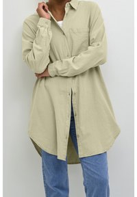 Chemise boutonnée vert clair avec une poche poitrine, manches longues et un ourlet arrondi, associée à un jean bleu. Texture de tissu douce.