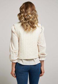Femme blonde aux cheveux ondulés portant un gilet matelassé crème sur une blouse à manches longues avec des poignets froncés et un jean bleu, de dos.