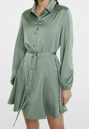 Kvinde iført lysegrøn satin skjortekjole med lange ærmer, knapper foran og bælte bundet i taljen mod ensfarvet baggrund.