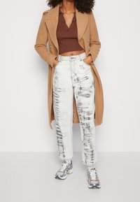 Brun kortärmad polo, krämfärgad och grå tie-dye vida jeans, och silverskor under en lång beige kappa. Mjuka material, avslappnad passform.