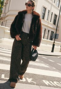 Braune Kunstpelzjacke, weißes T-Shirt, braune weit geschnittene Hosen mit Leopardenmuster, beige Plateau-Schuhe und eine schwarze gewebte Handtasche. Sonnige Straßenszene.