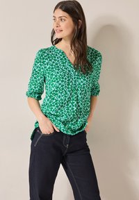 Grönmönstrad blus med V-ringning, långärmad som är uppvikt, gjord av lättviktig tyg. Parat med mörka jeans med ljusare sömmar.