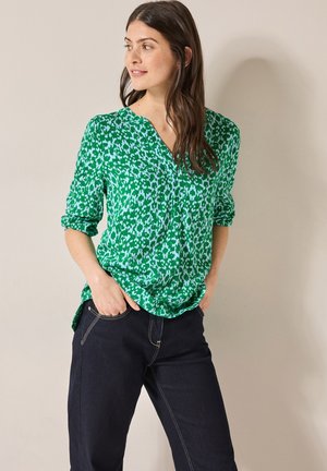 Groene gebloemde blouse met een V-hals, lange mouwen opgerold, gemaakt van lichte stof. Gecombineerd met donkere jeans met lichtere stiksels.