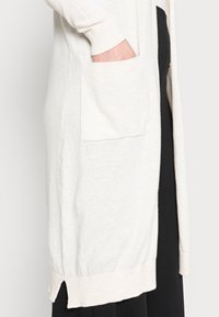 Dlouhý, světle béžový kardigán vyrobený z měkké tkaniny, s žebrovanými manžetami, bočním rozparkem na spodním lemu a nakládanou kapsou na levé straně.