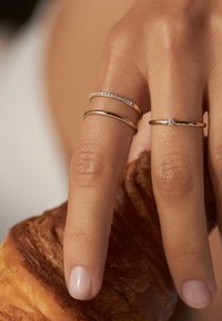 Drei Ringe aus Gold und Silber, einer mit kleinen Steinen besetzt, getragen an einem Finger, der ein Croissant hält, mit sichtbarem natürlichem Nagellack.