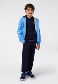 Giacca leggera blu sopra un hoodie blu navy, abbinata a pantaloni blu navy e sneaker nere con accenti blu. Design semplice, vestibilità rilassata.