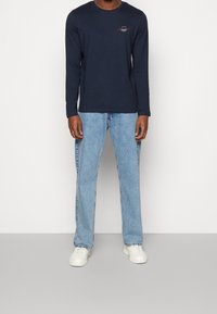 Camisa de manga larga azul marino con un pequeño logo rojo y blanco; combinada con jeans rectos de un azul claro y zapatillas blancas. Textura de tela suave.