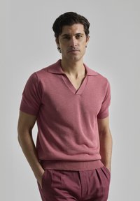Polo de manga corta y cuello en V en un material de punto rosa, con un collar y puños acanalados. Combinado con pantalones ajustados a juego.