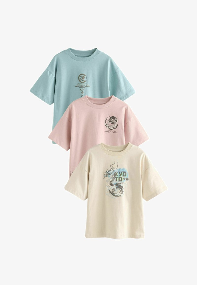 Tři nadměrná trička v pastelově modré, růžové a béžové barvě. Každé z nich má jedinečné grafiky: draka a názvy měst v kontrastních barvách.
