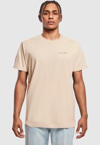 Beige cotton t-shirt featuring short sleeves and a crew neckline. Small green "DYSTOPIA" logo on the left chest. Relaxed fit.