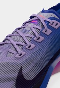 Κοντινό πλάνο μοβ και μπλε αθλητικού παπουτσιού με υφή από ύφασμα και σφιχτά δεμένα επίπεδα παστέλ λιλά κορδόνια πάνω από μία ραβδωτή γλώσσα.